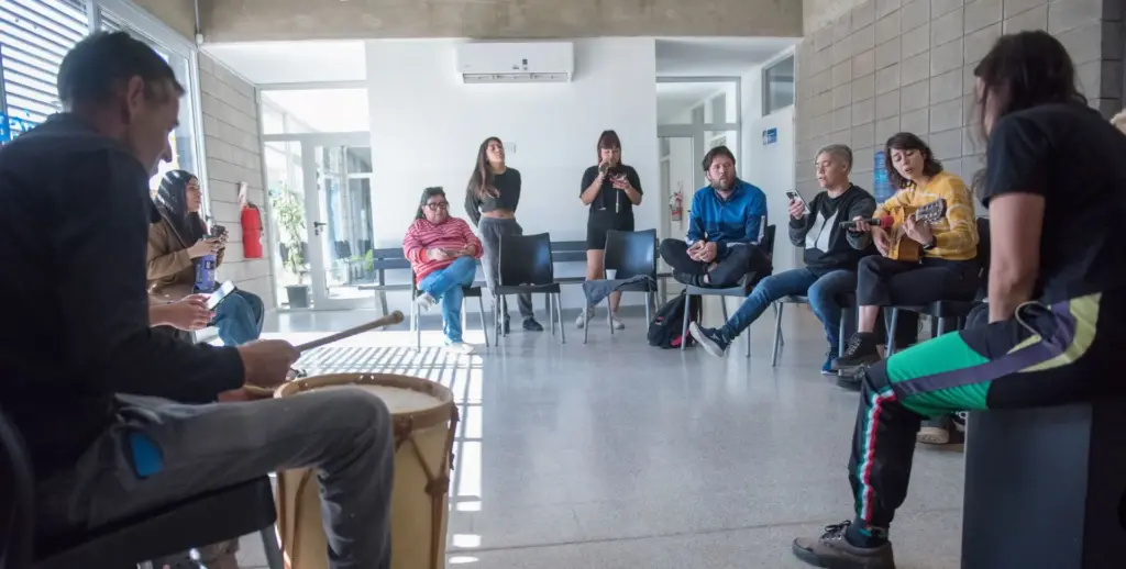 Grupo de personas sentadas en círculo en una sala luminosa del centro Raíces durante un taller de música, con un tambor y una guitarra