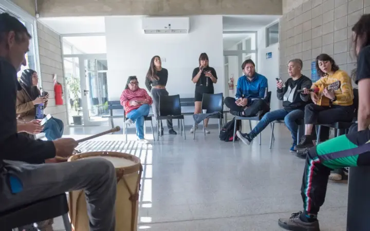 Grupo de personas sentadas en círculo en una sala luminosa del centro Raíces durante un taller de música, con un tambor y una guitarra
