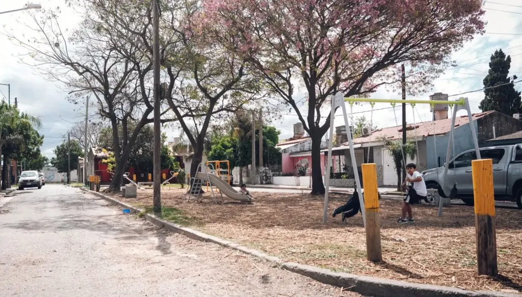 Calle de barrio con una plaza recién parquizada, árboles en flor, juegos infantiles y postes de madera en el borde de la vereda