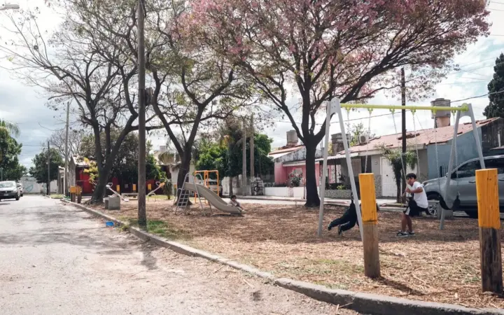 Calle de barrio con una plaza recién parquizada, árboles en flor, juegos infantiles y postes de madera en el borde de la vereda