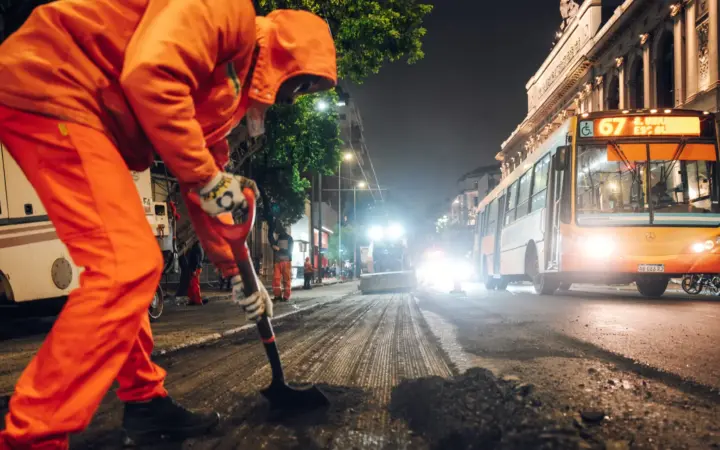 Operario con ropa naranja palea asfalto de noche en una calle de la ciudad, mientras un colectivo urbano circula junto a una máquina de obra
