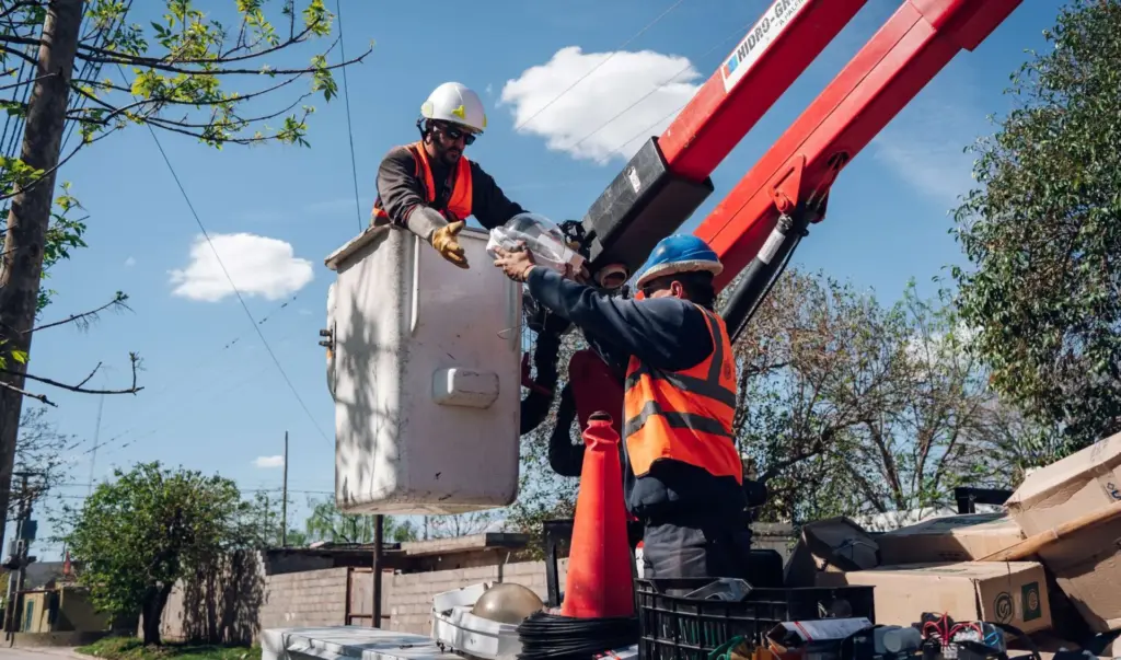 Dos operarios con casco y chaleco trabajan en la canasta elevada de un camión grúa para reemplazar una luminaria del alumbrado público
