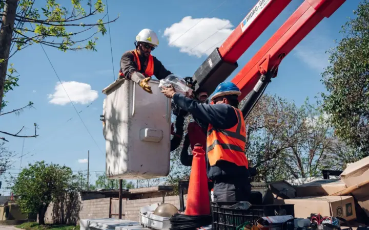 Dos operarios con casco y chaleco trabajan en la canasta elevada de un camión grúa para reemplazar una luminaria del alumbrado público