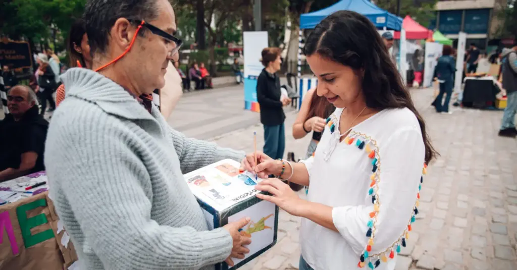 Una mujer y un hombre junto a una urna en un stand al aire libre durante una jornada de salud en la plaza