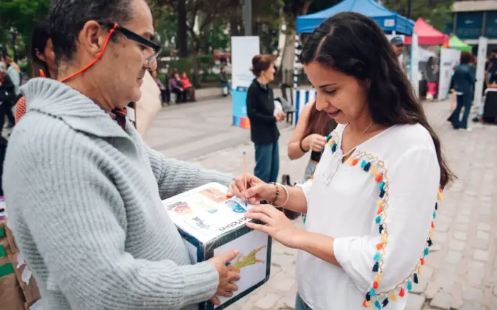 Una mujer y un hombre junto a una urna en un stand al aire libre durante una jornada de salud en la plaza